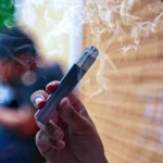 Person holding a silver vape pen emitting white vapor.
