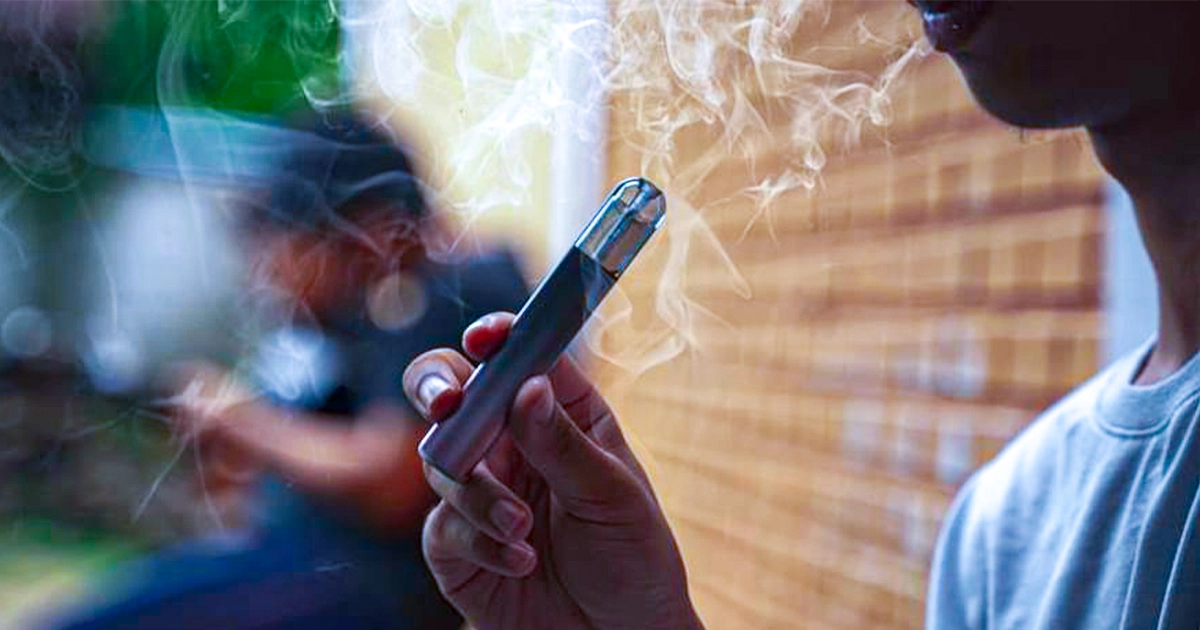 Person holding a silver vape pen emitting white vapor.