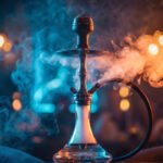 A glass and metal hookah sitting on a table, engulfed in thick, swirling smoke and illuminated by dramatic contrasting blue and orange lights.