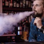 Bearded man exhaling a large cloud of vapor from a vape device in a bar setting.