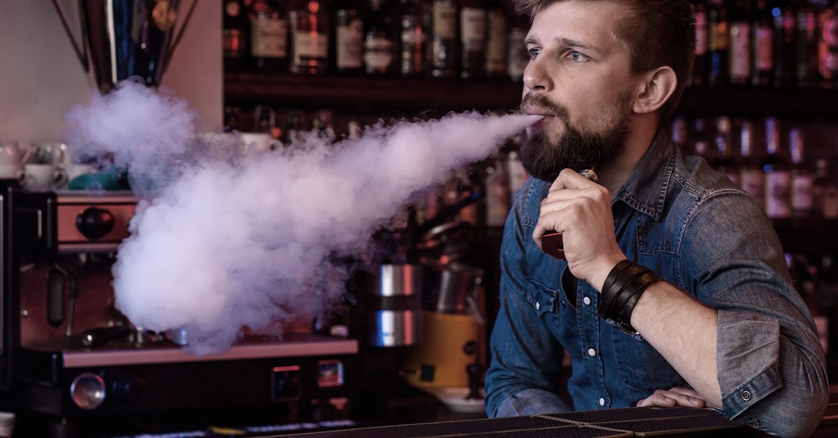 Bearded man exhaling a large cloud of vapor from a vape device in a bar setting.